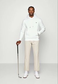Biely bavlnený mikina s predným vreckom a logom, béžové nohavice a biele golfové topánky. Drží golfovú palicu s čiernou rukoväťou.