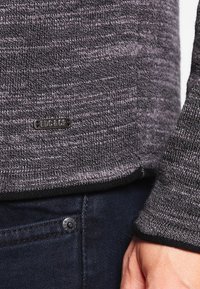 Pull en tricot gris foncé avec un motif subtilement texturé ; présente une étiquette en métal portant "EDG & CO" à l'ourlet. Associé à un jean foncé.