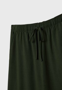Pantalon vert foncé avec ceinture élastique et cordon en tissu noué en nœud, présenté sur un fond uni gris clair.
