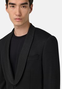 Svart kavaj med en glänsande svart slag, strukturerade axlar och en bröstficka; bärs över en svart rundhalsad tröja.