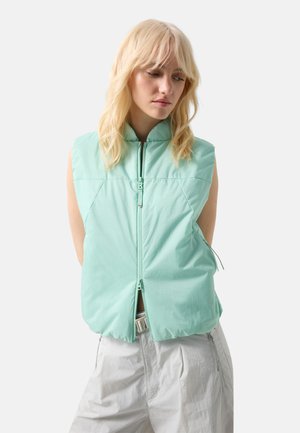 Blonde woman wearing a light turquoise padded sleeveless vest unzipped at the bottom and loose white pants against a plain background.