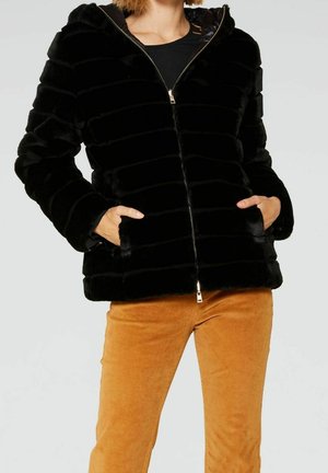 Kvinde iført en sort imiteret pelsjakke med lynlås og lommer samt sennepsfarvede kordfløjlsbukser, hænderne i jakkelommerne.