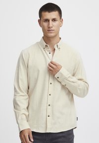 Chemise beige clair à manches longues avec col à boutons, présentant des boutons foncés, un tissu texturé et un ourlet légèrement courbé.
