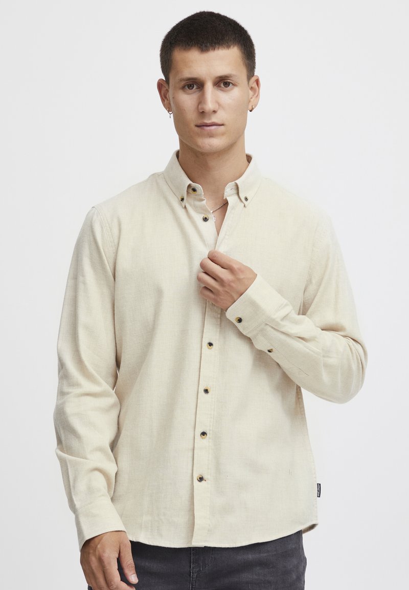 Chemise beige clair à manches longues avec col à boutons, présentant des boutons foncés, un tissu texturé et un ourlet légèrement courbé.