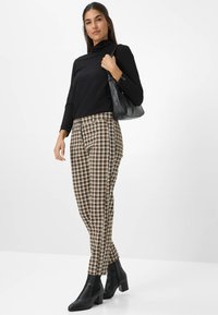 Zwarte lange mouw turtleneck, geblokte broek in bruin, wit en zwart. Model draagt zwarte enkellaarsjes en houdt een zwarte handtas vast.