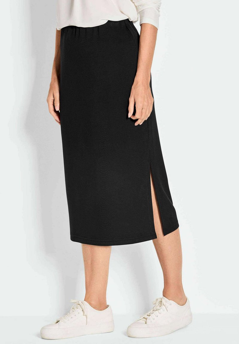 Jupe noire en tissu doux, longueur mi-mollet, avec une fente latérale pour faciliter le mouvement. Associée à des baskets blanches.