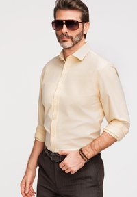 Homme avec des lunettes de soleil foncées, chemise beige boutonnée avec manches retroussées, pantalon sombre et bracelets au poignet sur fond uni.