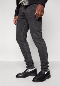 Vaqueros de mezclilla negra con costuras blancas contrastantes, corte ajustado y bolsillos, combinados con mocasines negros brillantes y calcetines grises.