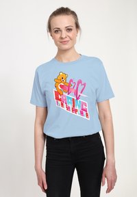 Světle modré bavlněné tričko s potiskem kreslené medvěda a textem v sytých barvách. Krátké rukávy a volný střih. Černé džíny pod ním.