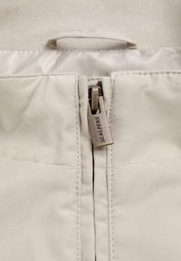 Tejido beige con una textura suave que presenta una cremallera visible, un tirador de metal con la marca "SCALPERS" y pliegues sutiles en la parte superior.