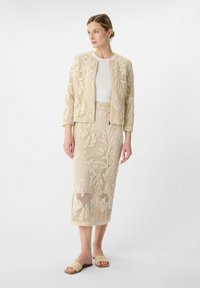 Beżowy zestaw z koronkową marynarką i spódnicą w kwiatowe wzory, zestawiony z białą bluzką i beżowymi sandałami. Teksturowany, dopasowany krój.