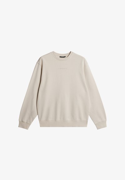 Beige pulóver, kerek nyakkivágással, hosszú ujjaival és bordázott mandzsettával. Középen egy diszkrét logó található. Simább textúrájú és lezser dizájnnal rendelkezik.