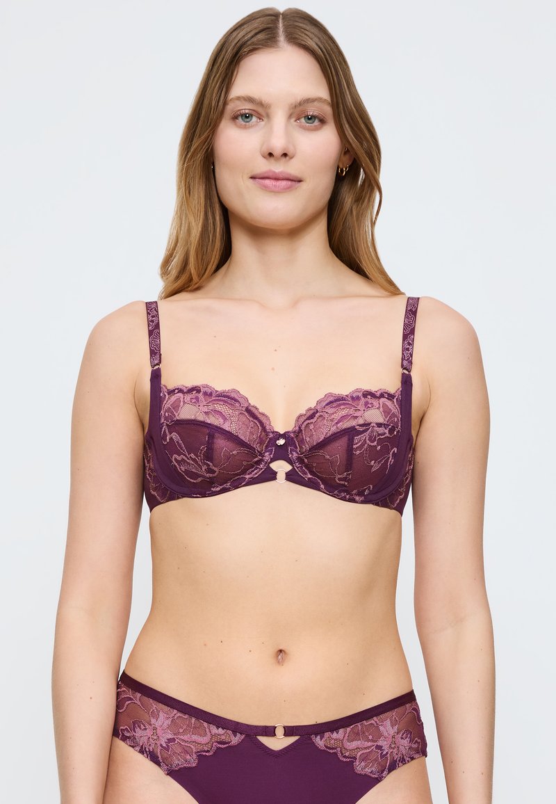 Krajkovaná fialová podprsenka s kosticemi, průhlednými košíčky a nastavitelnými ramínky. Obsahuje květinové vzory a centrální ozdobný kovový detail.