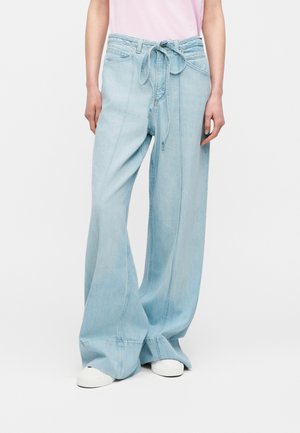 Άτομο που φοράει ανοιχτό μπλε φαρδιά τζιν παντελόνια με δεμένη μέση και λευκά αθλητικά παπούτσια, στέκεται μπροστά σε απλό φόντο.