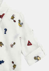 Camicia bianca con disegni ricamati colorati, inclusi simboli, punti interrogativi e forme. Presenta un polsino e un colletto con bottoni.