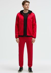 Chándal rojo con chaqueta con cremallera y pantalones, detalles en negro, bolsillos con cremallera y texto impreso en la chaqueta. Tejido texturizado y ajuste slim.