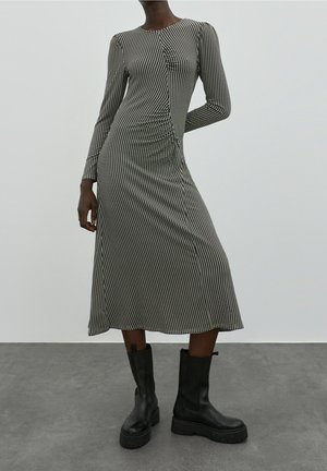 Robe en maille à carreaux noir et blanc, manches longues, col rond, et coupe mi-longue. Portée avec des bottes noires épaisses.