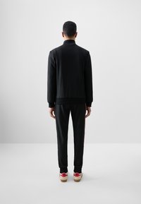 Black athletic jacket and pants, soft fabric, featuring ribbed cuffs, and contrasting red side stripes. Footwear has pink accents and woodgrain soles.