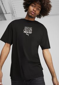 Juodas medvilninis marškinėlis su laisvu kirpimu, su baltu tekstu "HOUSE & GROOVE" ir logotipu krūtinėje. Trumpi rankovės ir apvalus kaklo iškirpimas.