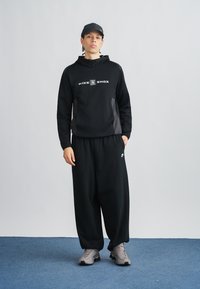 Μαύρο φούτερ Nike Shox με λογότυπο, αντίθετες γκρι λεπτομέρειες, ταιριαστό μαύρο παντελόνι φόρμας και γκρι αθλητικά παπούτσια σε μπλε φόντο μοκέτας.