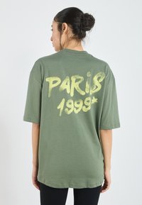 Grøn oversized T-shirt lavet af bomuld med fed gul tekst "PARIS 1999" på ryggen. Korte ærmer og design med rund hals.