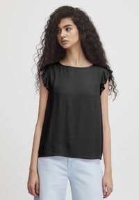 Top nero senza maniche con dettagli arricciati, tessuto texturizzato, scollatura rotonda, vestibilità ampia, indossato con jeans azzurri chiari, design minimalista.