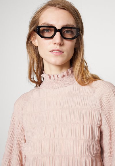 Μαύρα ορθογώνια γυαλιά και ένα ανοιχτό ροζ, υφασμάτινο φόρεμα με ριχτό ψηλό γιακά και κατακόρυφες πτυχές, διαθέτοντας μακριά μανίκια.