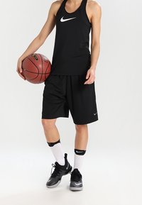 Schwarzes Nike Basketball-Top und Shorts, kombiniert mit schwarz-grauen Turnschuhen. Weiße Socken mit schwarzen Akzenten. Hält einen Basketball.