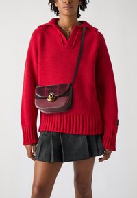 Maglione rosso a maglia con orlo e colletto a coste, abbinato a una gonna a pieghe nera a quadretti. Una piccola borsa a tracolla color bordeaux presenta una chiusura dorata.