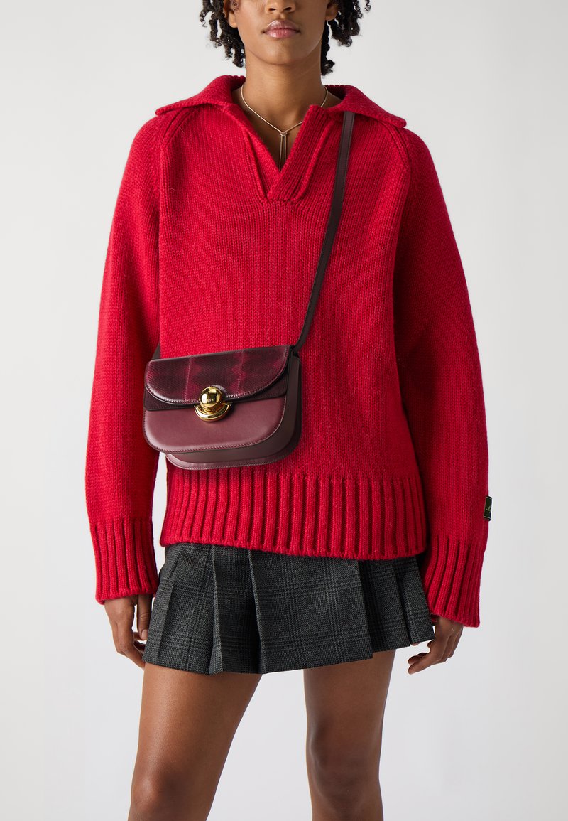 Maglione rosso a maglia con orlo e colletto a coste, abbinato a una gonna a pieghe nera a quadretti. Una piccola borsa a tracolla color bordeaux presenta una chiusura dorata.