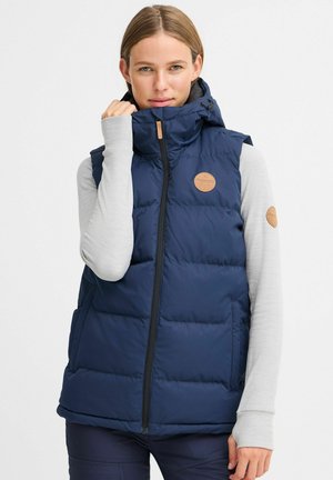 Gilet rembourré bleu marine avec un col haut, fermeture éclair et poches latérales. Présente un patch logo en cuir marron sur la poitrine.