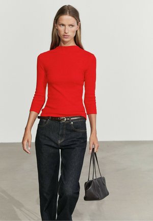 Femme portant un haut moulant rouge vif et un jean foncé tenant un petit sac à main noir sur un fond clair uni.