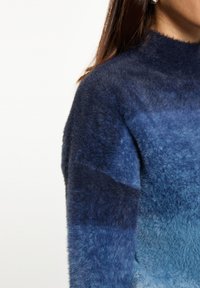Pullover morbido nelle tonalità di blu con colletto alto. Presenta un motivo a gradiente e una texture morbida, offrendo un aspetto accogliente.