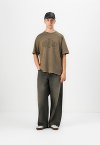Ungdommelig mann iført en overdimensjonert brun t-skjorte med grafikk, løse mørke jeans, hvite joggesko og svart baseballcaps som står mot en hvit bakgrunn.
