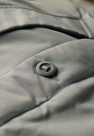Tessuto grigio chiaro, morbido, con una finitura liscia, caratterizzato da un bottone rotondo con la scritta "SUPERDRY" in rilievo. La cucitura testurizzata evidenzia il design.