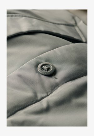 Tessuto grigio chiaro, morbido, con una finitura liscia, caratterizzato da un bottone rotondo con la scritta "SUPERDRY" in rilievo. La cucitura testurizzata evidenzia il design.