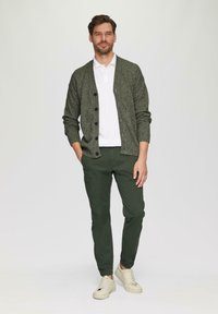 Grüner strukturierter Cardigan mit schwarzen Knöpfen, getragen über einem weißen Poloshirt. Kombiniert mit dunkelgrünen Hosen und hellen Sneakers.
