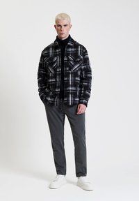 Camicia a quadri bianco e nero con doppie tasche sul petto, abbinata a pantaloni grigi e sneakers bianche. Texture liscia, design strutturato, vestibilità moderna.