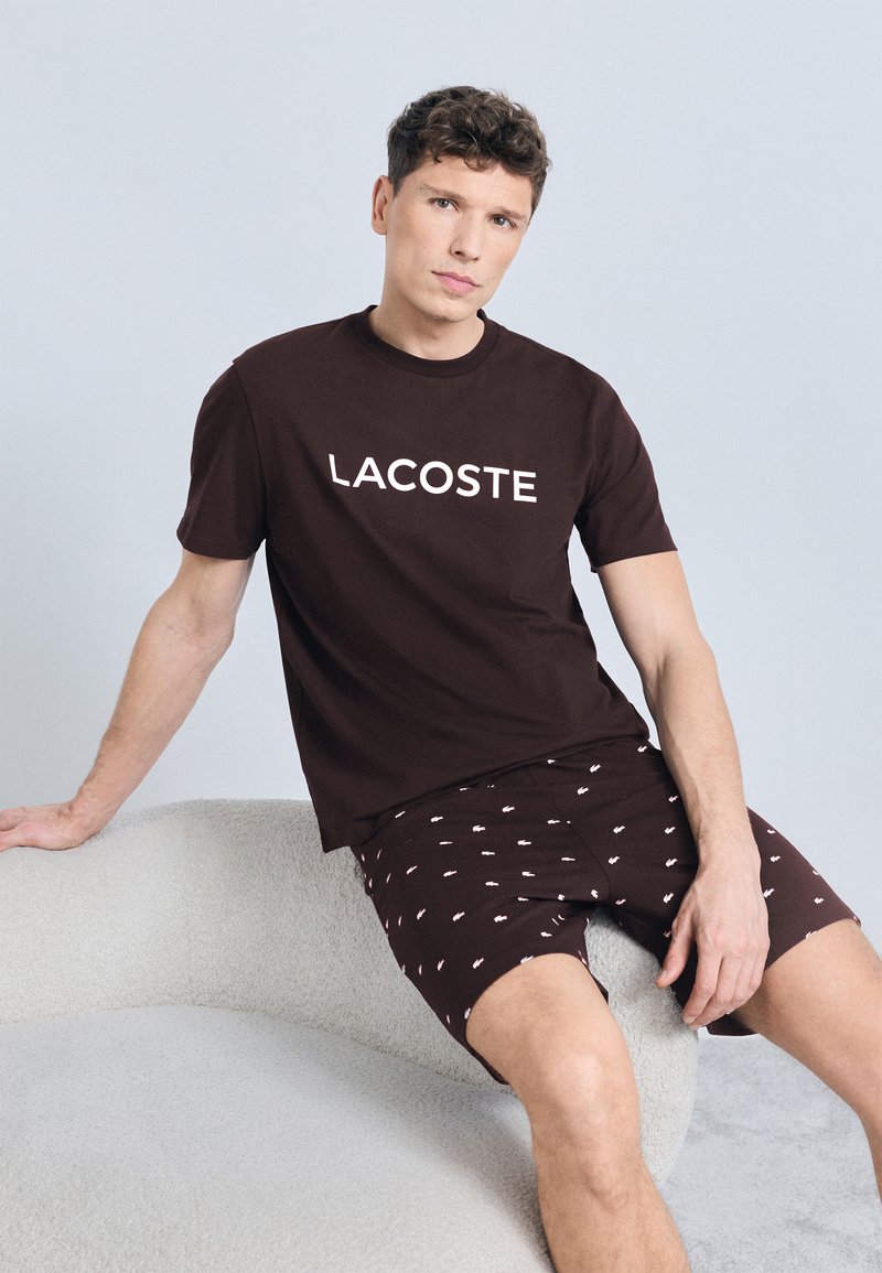 Άνδρας που φοράει καφέ t-shirt Lacoste και σορτς που ταιριάζουν, καθισμένος σε έναν ανοιχτόχρωμο καναπέ με υφή, μπροστά σε απλό φόντο.