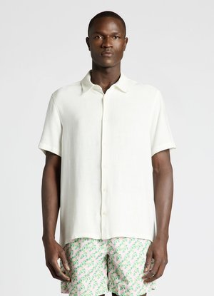 Homme portant une chemise blanche à manches courtes boutonnée et un short rose avec un motif de feuilles vertes, debout devant un fond uni.