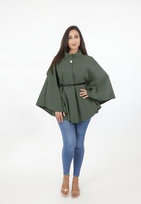 Grön, strukturerad cape med hög krage och en enda knapp, åtdragen i midjan med ett svart bälte. Bärs över blå, åtsittande jeans.