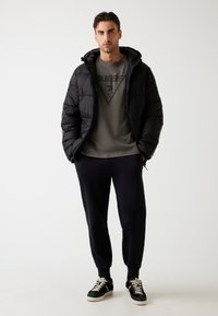 Hombre vestido con una chaqueta de abrigo negra, camiseta gris con logo, pantalones de chándal negros y zapatillas negras con cordones blancos, está de pie con las manos en los bolsillos.