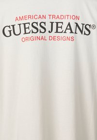 Fehér, pamut póló, amelyen fekete és piros felirat található: "AMERICAN TRADITION GUESS JEANS® EREDETI TERVEZÉSEK".