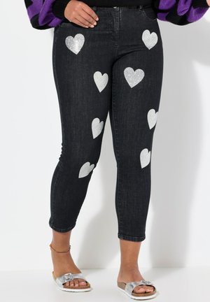 Zwarte jeans met glinsterende zilveren hartpatronen verspreid over de benen. Strakke pasvorm met een enkel-lange zoom, gecombineerd met zilveren sandalen.