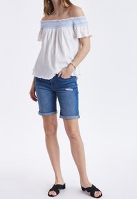 Top bianco con spalle scoperte, dettagli in smocking blu e maniche a volant, abbinato a pantaloni denim corti arrotolati. Sandali a scivolo neri completano il look.