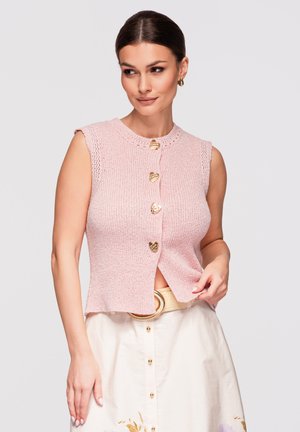 Vrouw draagt een mouwloos roze gebreid topje met gouden knopen in de vorm van een hart en een crèmekleurige rok met gouden riem en knopen voor een lichte achtergrond.