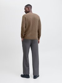 Bărbat în picioare cu mâinile în buzunare, purtând un pulover maro texturat, pantaloni gri și pantofi negri, cu fața spre spate, pe un fundal simplu.