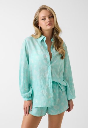 Blonde Frau trägt hellblau gemustertes langärmeliges Hemd und passende Shorts, steht vor einem schlichten weißen Hintergrund.