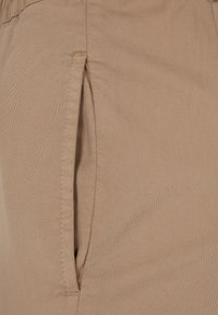 Pantalon beige en tissu lisse et texturé avec une poche latérale et des détails de couture subtils.