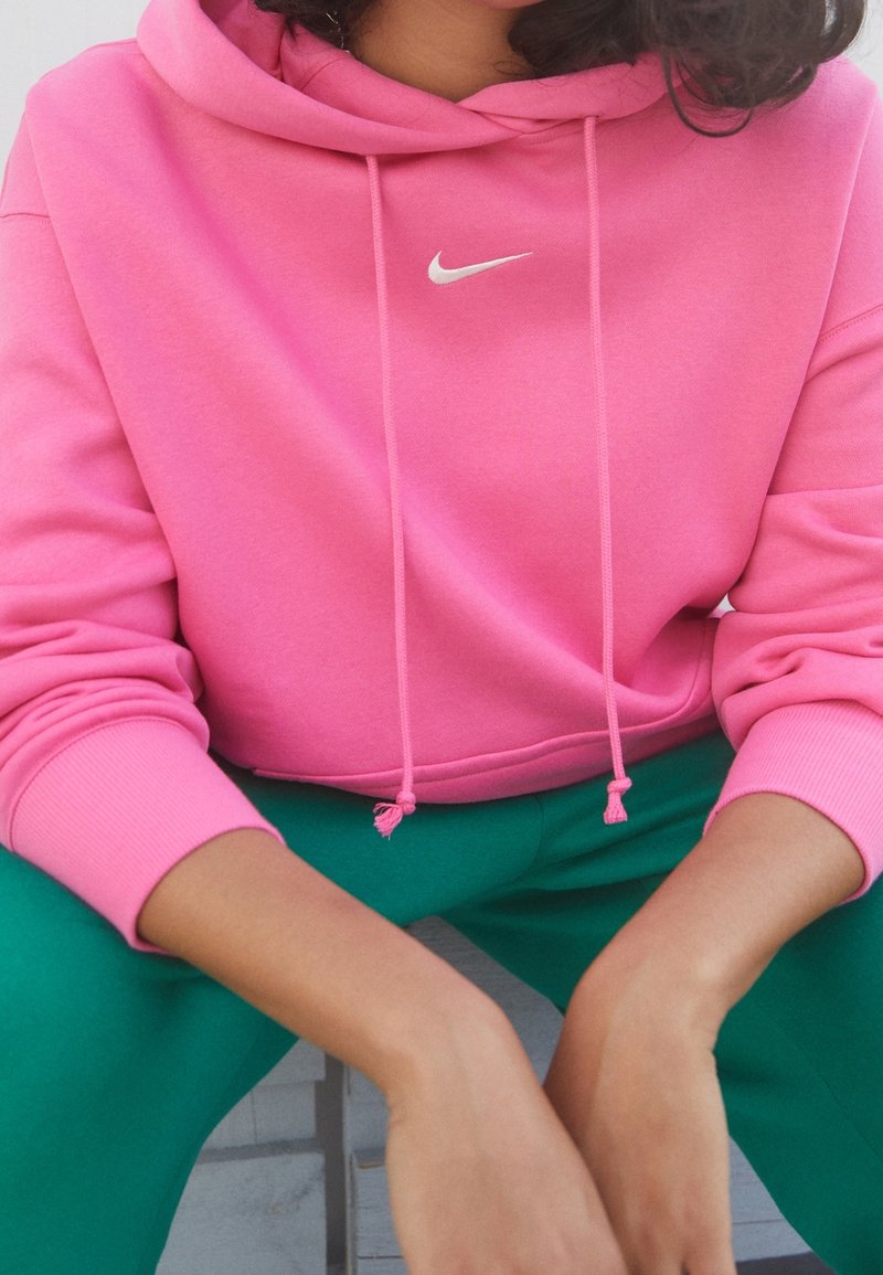Pink cropped hoodie with a white Nike logo, featuring drawstrings and ribbed cuffs. Paired with green pants, showing a relaxed fit.